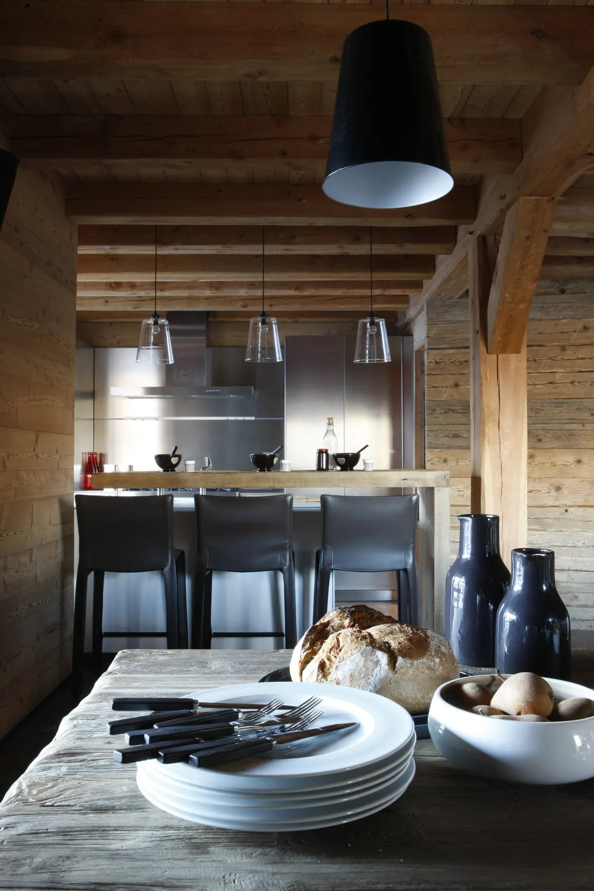 Grande table de bois dressée sous des suspensions noires, cuisine en arrière-plan