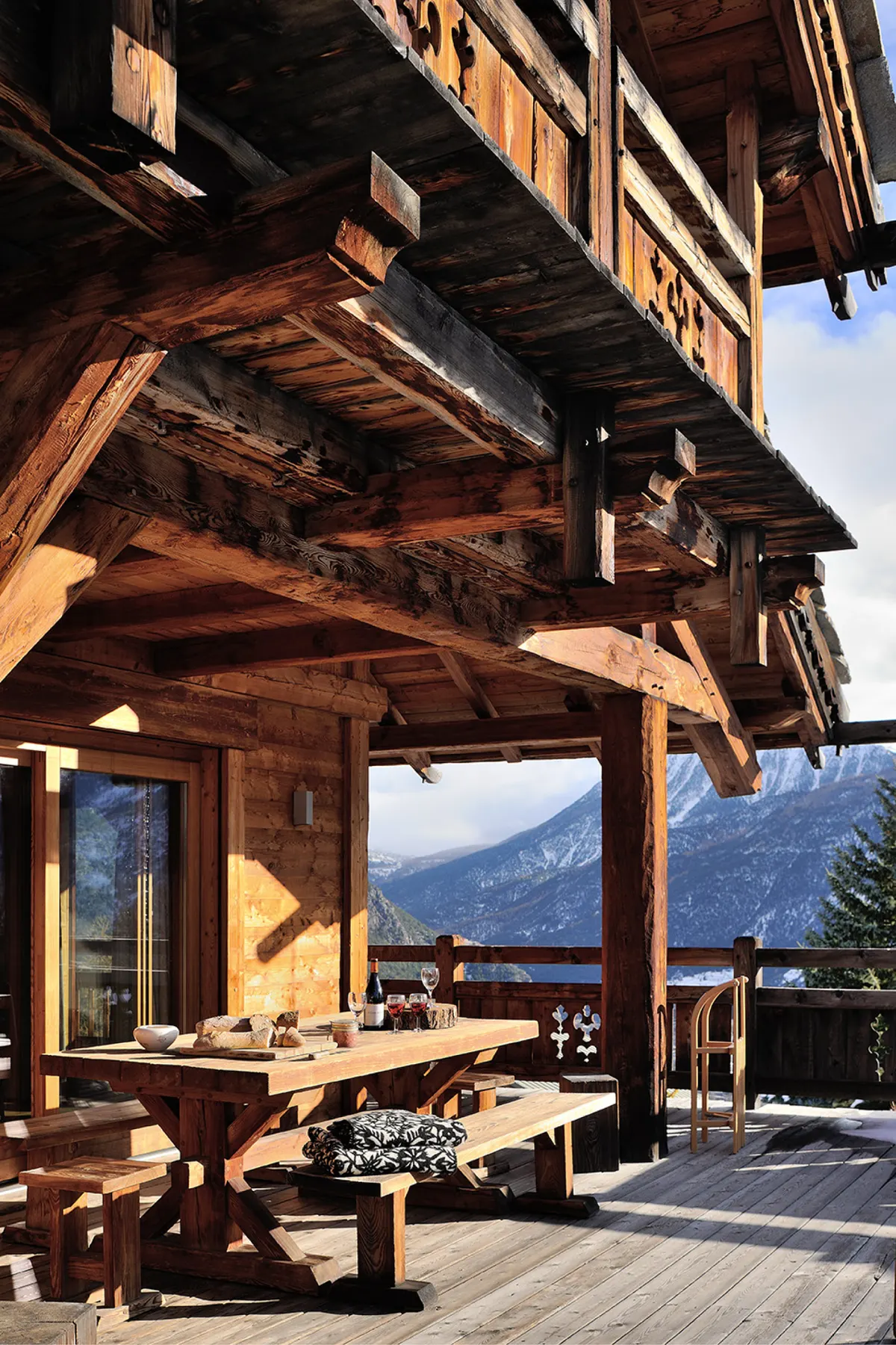 Terrasse couverte sous les avancées de toit en mélèze, grande table en bois face aux sommets enneigés